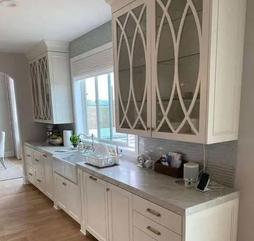 Custom white kitchen cabinets with decorative glass-front upper doors, a farmhouse sink, and tile backsplash