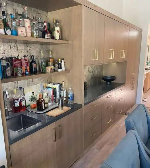 Custom wood-tone home bar with a black countertop, mosaic tile backsplash, gold hardware, and a bar sink