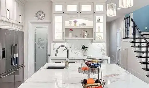 Custom white kitchen cabinets with marble countertops and backsplash, next to a pantry and staircase