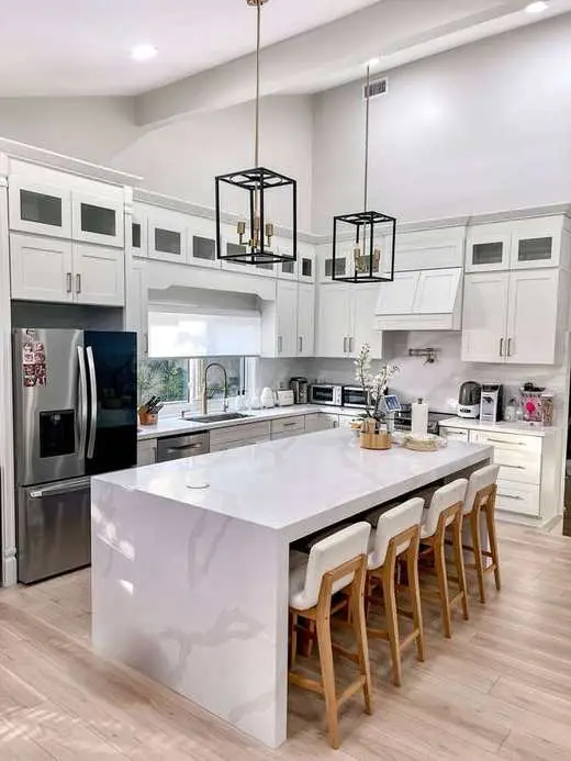 Wide view of a custom white kitchen with a marble waterfall island, vaulted ceiling, and black pendant lighting